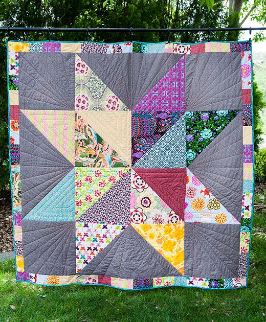 a patchwork quilt hanging on a clothesline in the grass with trees behind it