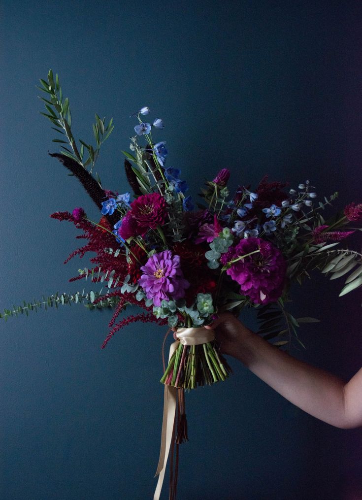 a woman holding a bouquet of flowers in front of a blue wall with purple and green foliage