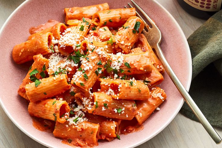 a pink bowl filled with pasta covered in sauce and parmesan cheese next to a fork