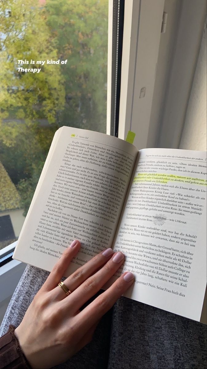 a woman is holding an open book in her hand and looking out the window at trees