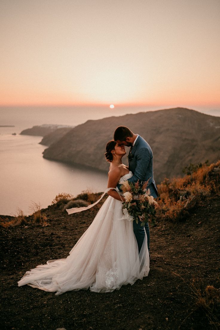 a bride and groom standing on top of a hill next to the ocean at sunset