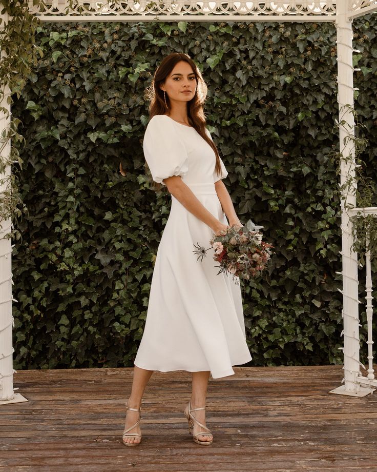 a woman in a white dress standing on a wooden floor with greenery behind her