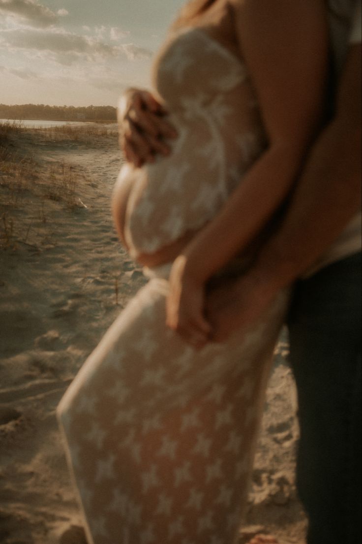 a pregnant woman standing on top of a sandy beach next to a man holding her belly