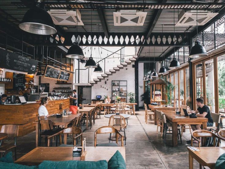 people sitting at tables in a restaurant with lots of windows and hanging lights above them