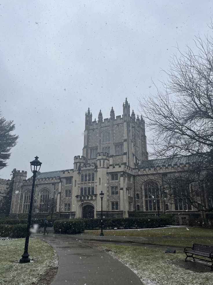 an old building with snow falling on the ground and benches in front of it,