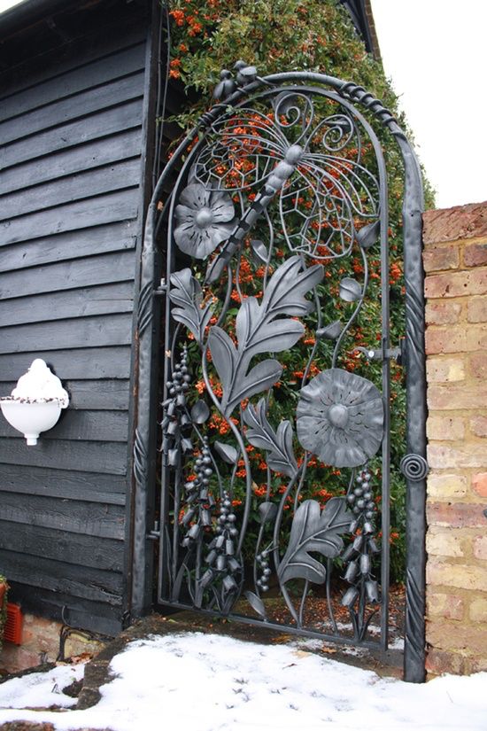 an iron gate with flowers and leaves on the side of a black building next to a brick wall