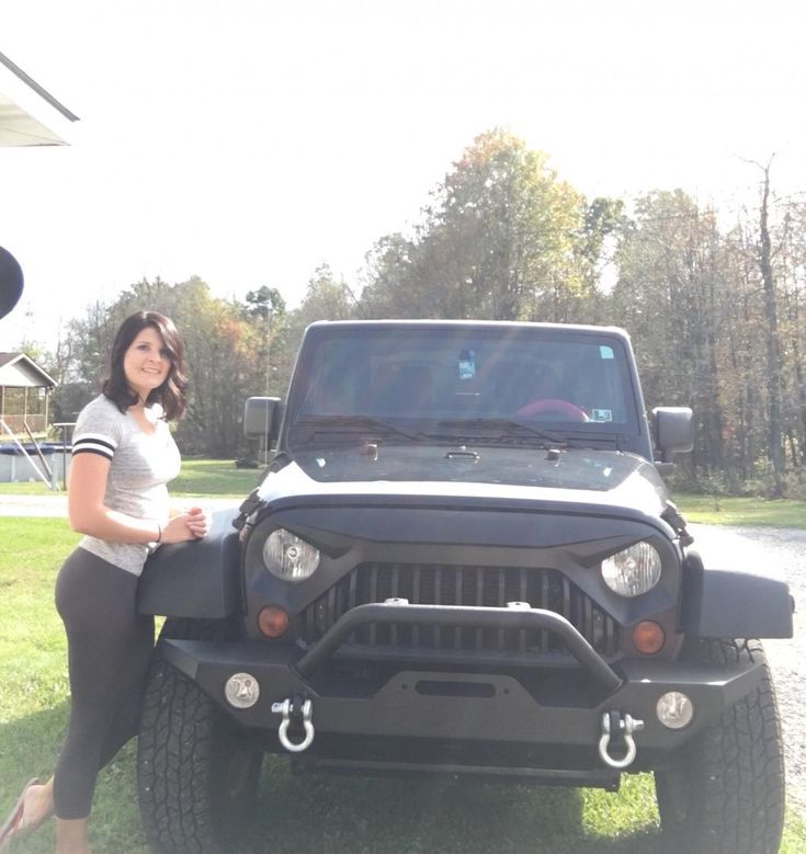 a woman sitting on the hood of a black jeep in front of a park with trees