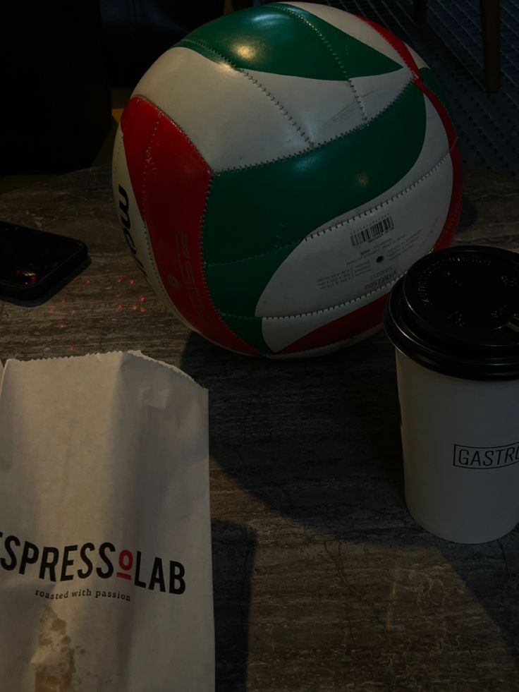 a ball, cup and cell phone sitting on a table next to a paper bag