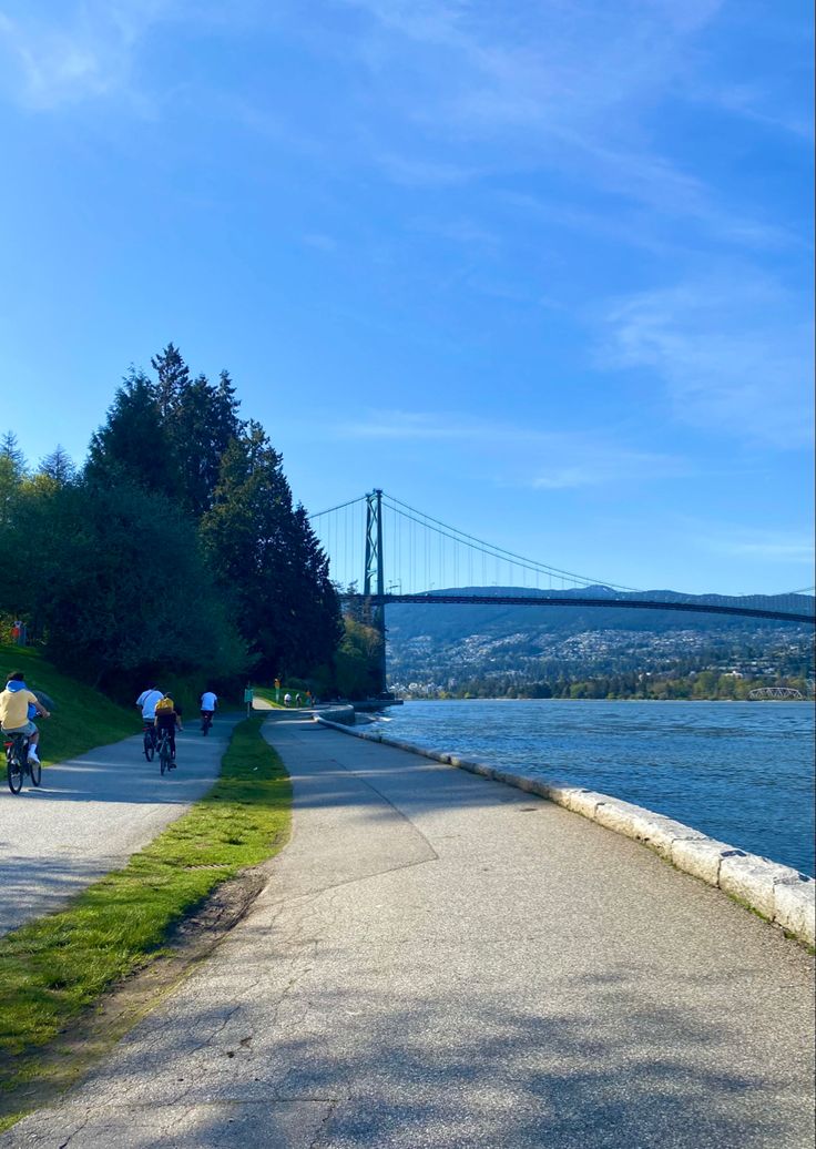 two people riding bikes on a paved path next to the water with a bridge in the background