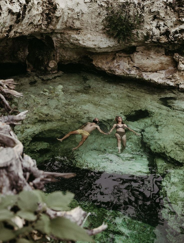 two people are floating in the water near some rocks and mossy vegetation, while holding hands