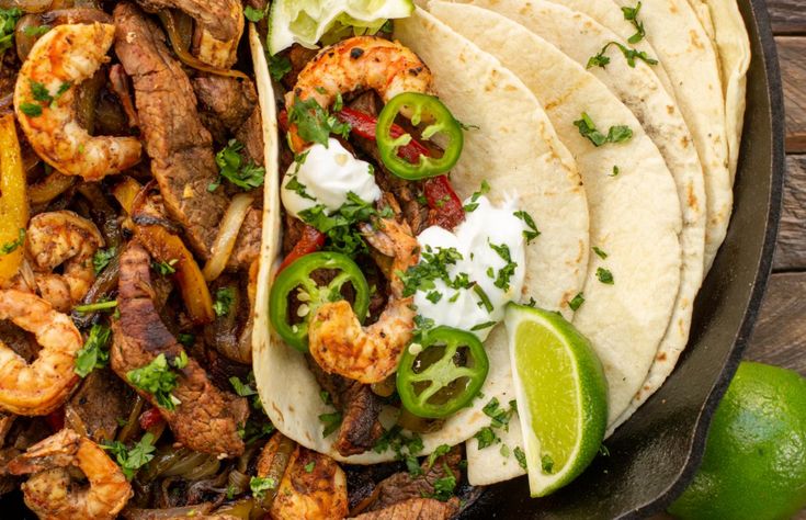 a skillet filled with shrimp tacos and tortillas next to lime wedges