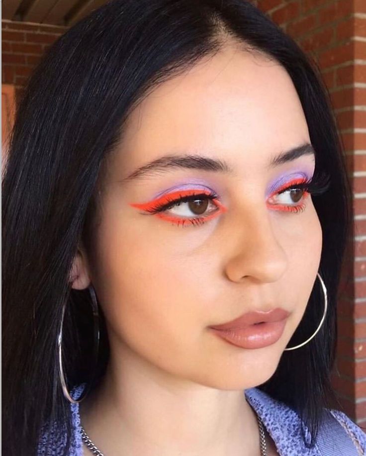 a woman with long black hair wearing bright orange and pink eye makeup looks at the camera