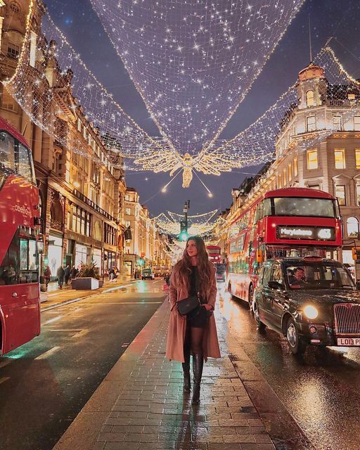 a woman is walking down the street in front of double decker buses and christmas lights