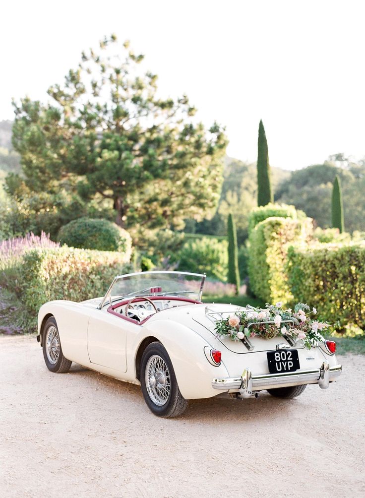 an old white sports car parked in front of some bushes and trees with flowers on the hood