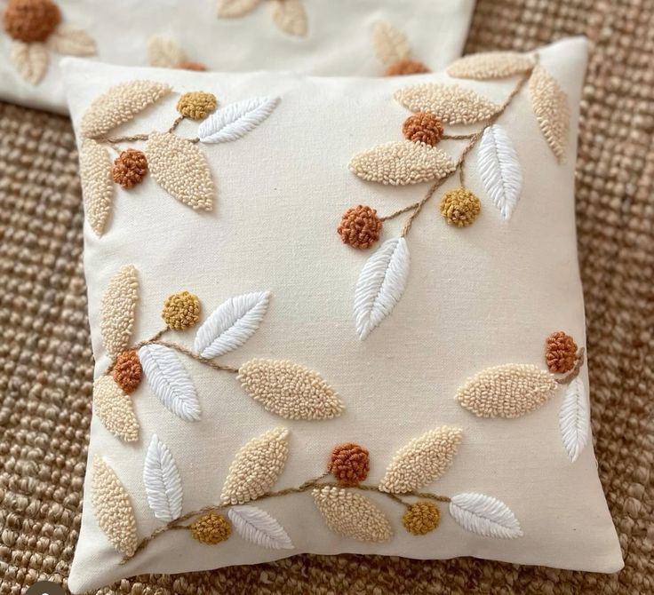 two white pillows with embroidered leaves on them sitting on a brown carpeted floor next to each other