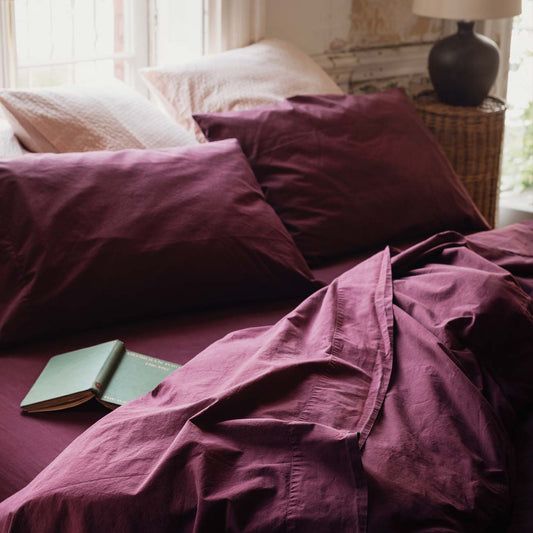 a bed with purple sheets, pillows and a book on it's side in front of a window