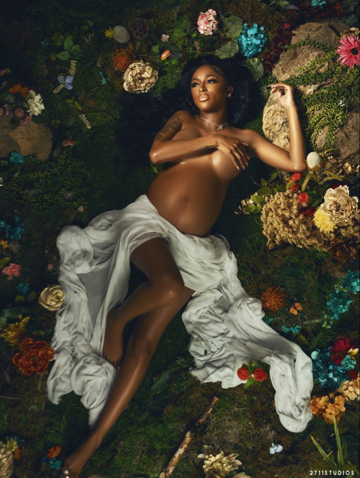 a woman laying on the ground surrounded by flowers and plants, with her body covered in flowing fabric