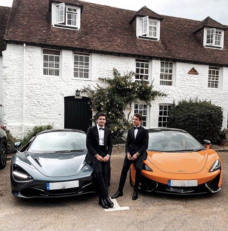 two men in tuxedos standing next to an orange sports car and a white house