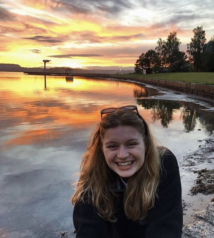 a young woman sitting on the edge of a body of water smiling at the camera