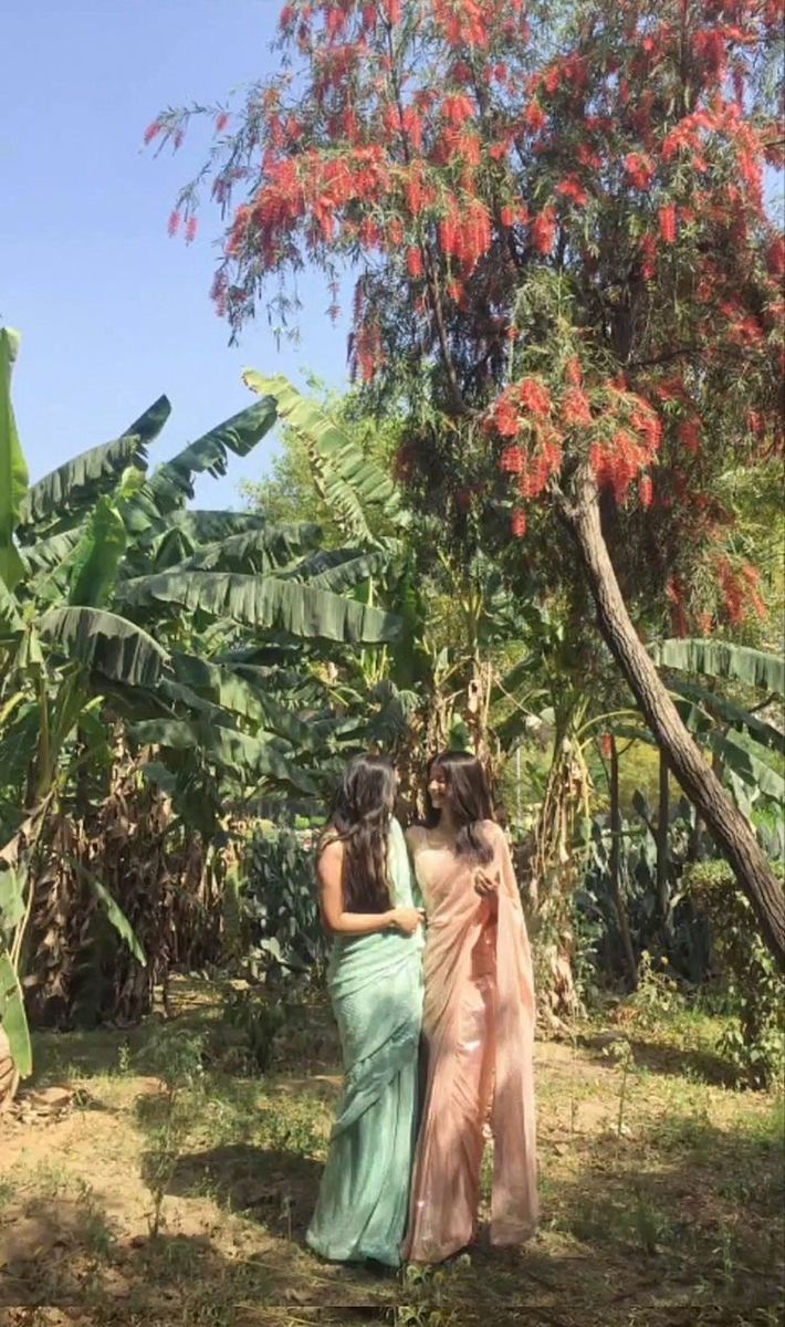 two women in sari standing next to each other near some trees and bushes with red flowers on them