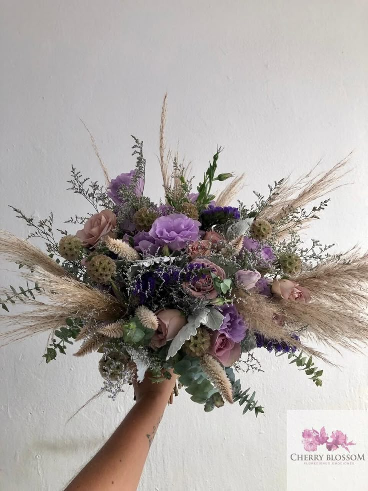 a hand holding a bouquet of purple flowers and grasses in front of a white wall