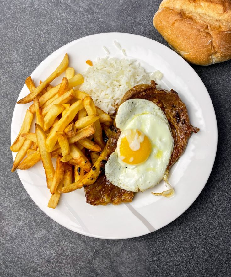 a white plate topped with meat, fries and an egg on top of rice next to a roll