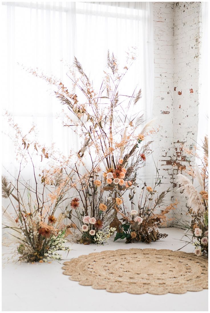 an arrangement of flowers and plants in front of a white brick wall with a round rug on the floor