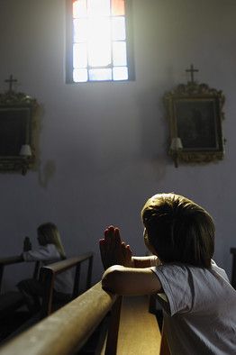 a young boy sitting at the end of a church pew with his hands clasped in front of him