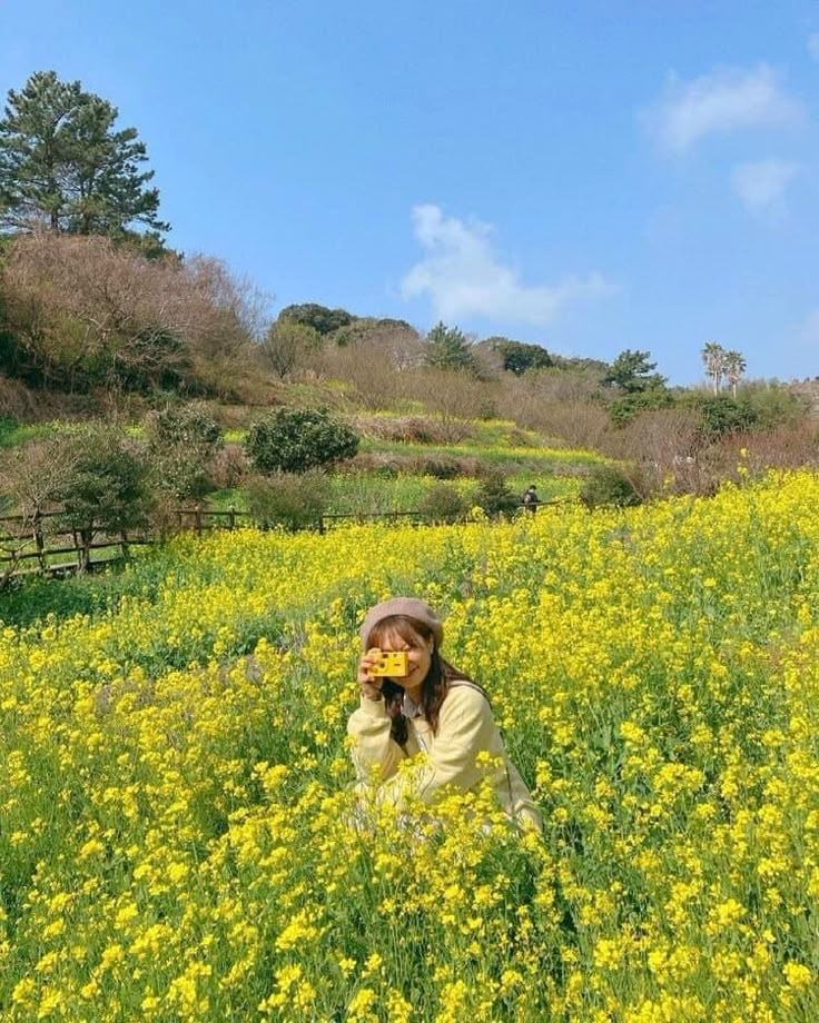 a woman is sitting in a field with yellow flowers and holding a camera up to her face