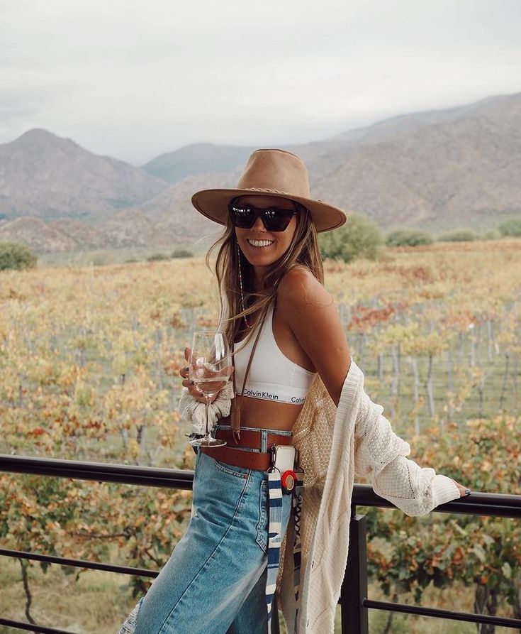 a woman standing in front of a vineyard holding a wine glass and smiling at the camera