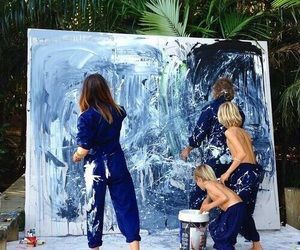 three women in blue jumps painting on a large piece of art with palm trees behind them