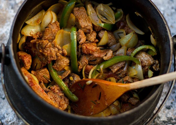 a pot filled with meat and vegetables on top of a stone counter next to a wooden spoon