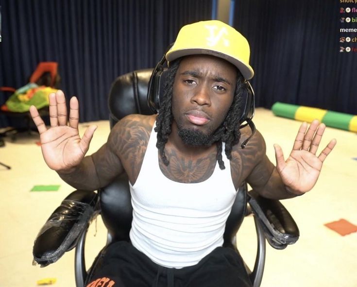 a man with dreadlocks sitting in a chair wearing a yellow hat and holding his hands up