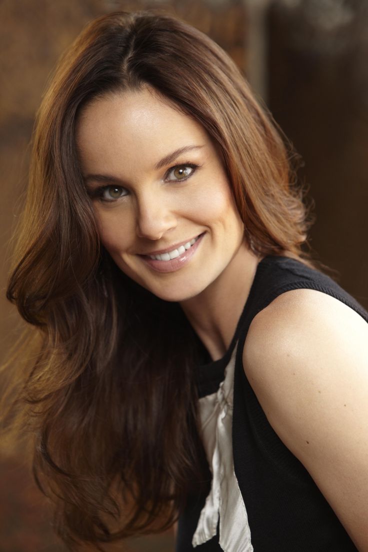 a woman with long brown hair smiling at the camera and wearing a black vest over her shoulders