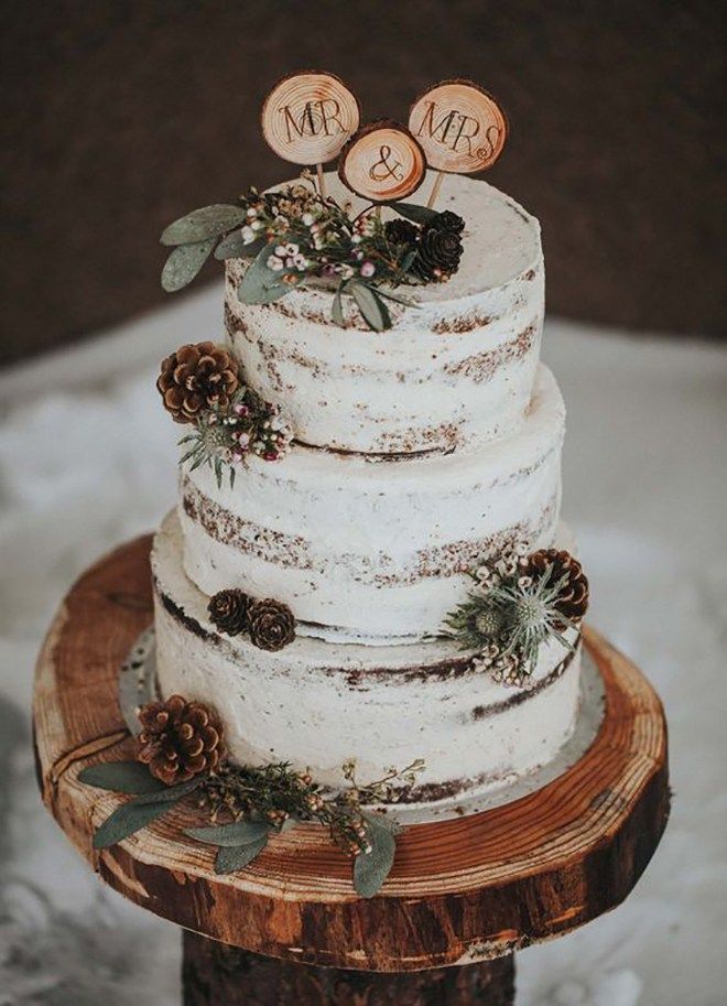 a white wedding cake with pine cones and greenery on top is sitting on a wooden stand