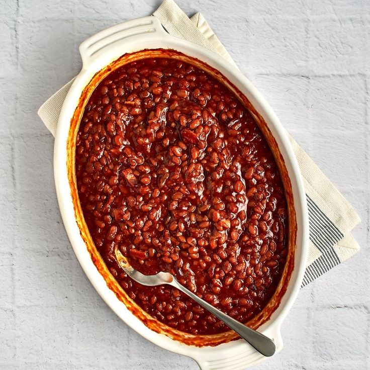 a bowl filled with baked beans on top of a white table cloth next to a fork