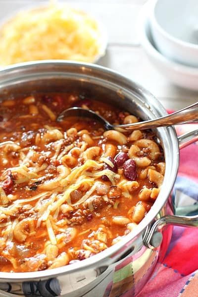 a large pot filled with chili and beans on top of a colorful table cloth next to silver spoons