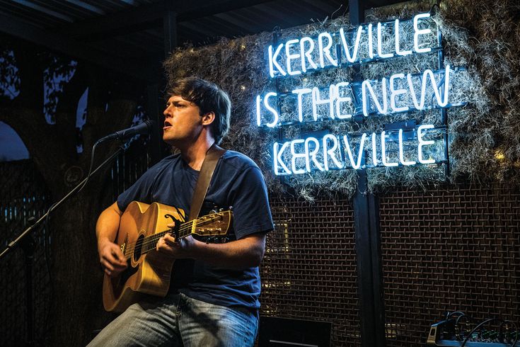 a man playing a guitar in front of a neon sign that reads kernvillee is the new kervillee