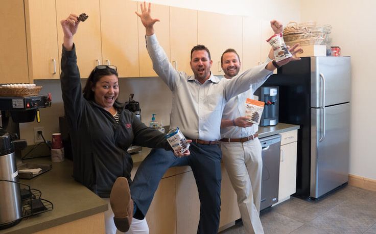 two men and a woman standing in a kitchen with their arms up, holding food