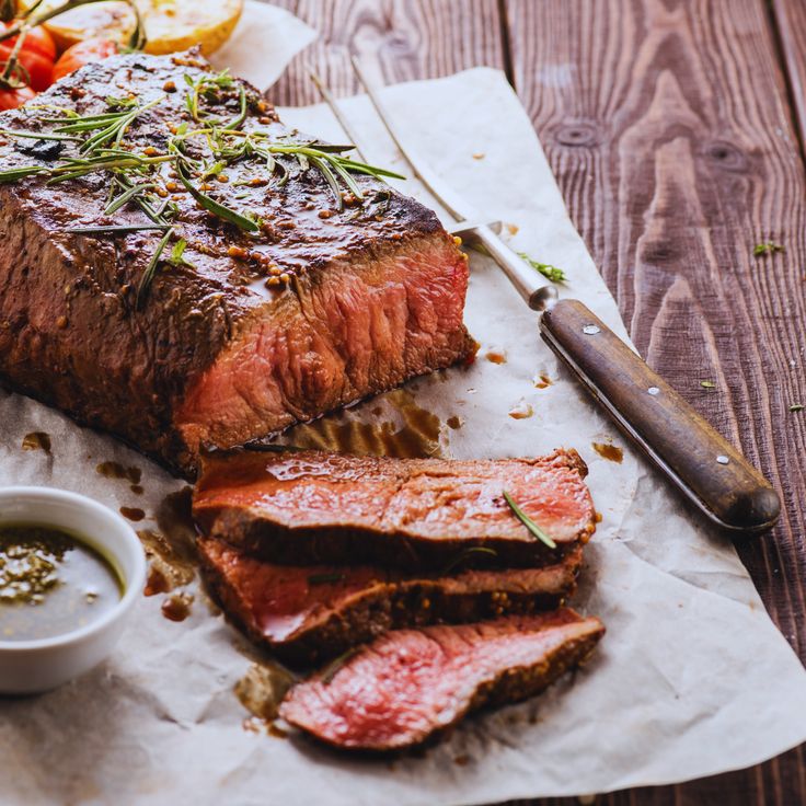 a piece of steak on a cutting board next to a knife and bowl of sauce