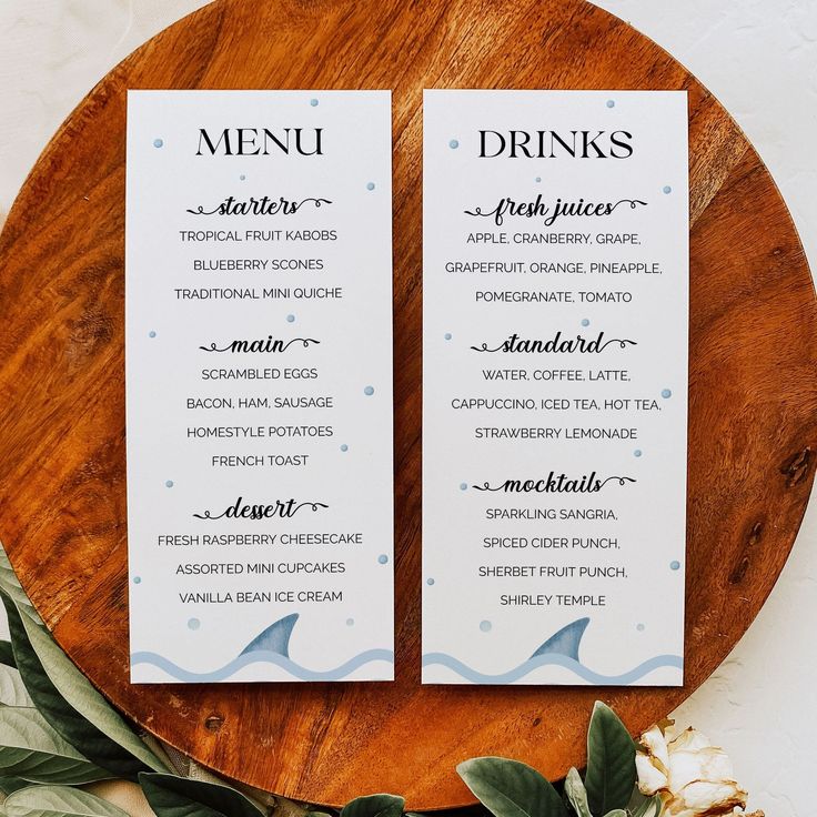 two white menu cards sitting on top of a wooden platter next to green leaves