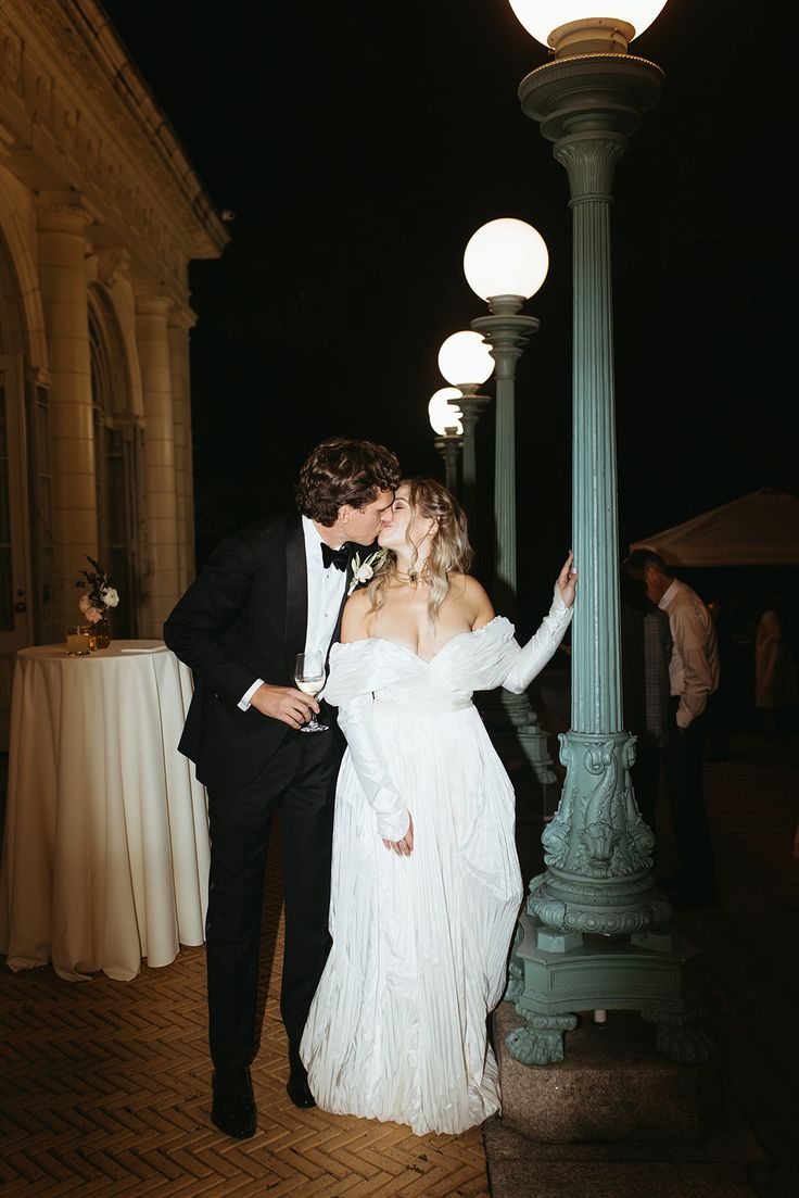 a bride and groom kissing in front of a street light at night with their arms around each other