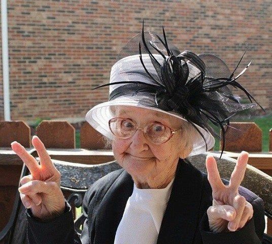 an old woman wearing a hat and glasses making the peace sign with her hands while sitting on a bench