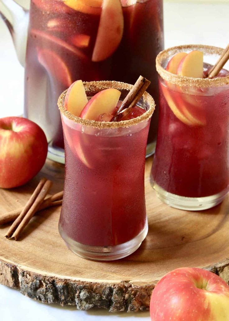 two glasses filled with apple cider on top of a cutting board next to apples