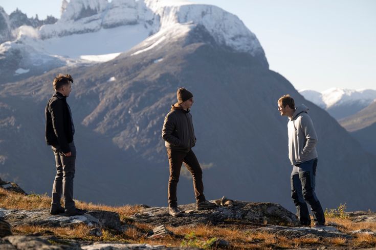 three young men standing on top of a rocky hill looking at the mountain range in the distance