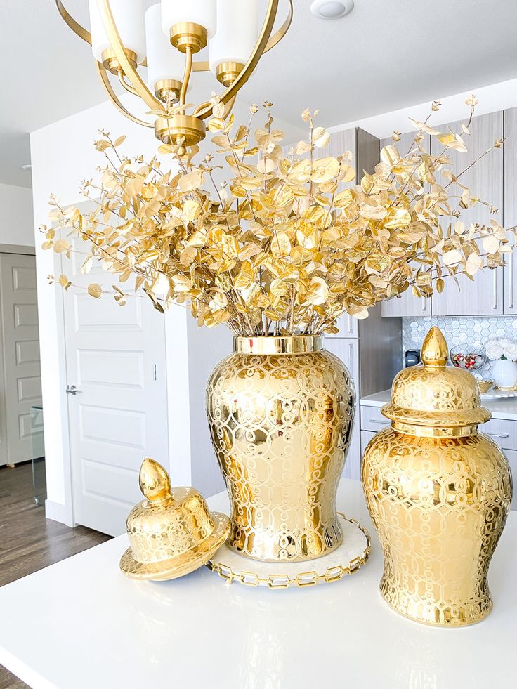 gold vases with flowers in them sitting on a white countertop next to a chandelier