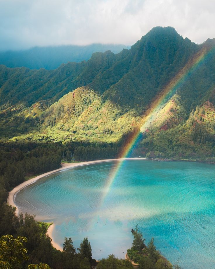 a rainbow shines in the sky over a blue lake and mountain range with green trees