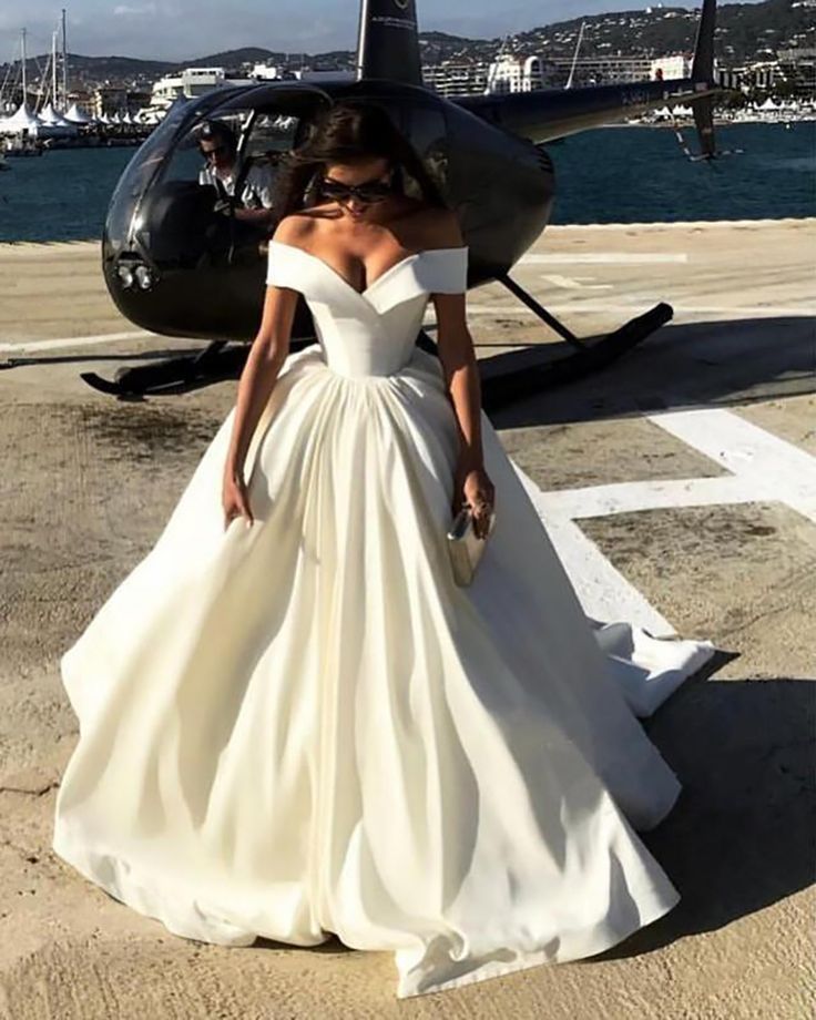 a woman standing in front of a helicopter wearing a wedding dress with off the shoulder sleeves