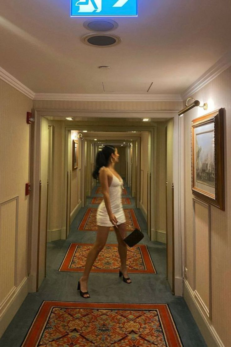 a woman in white dress walking down a hallway with an exit sign above her head