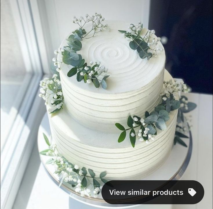 a white wedding cake decorated with greenery and flowers on a table next to a window